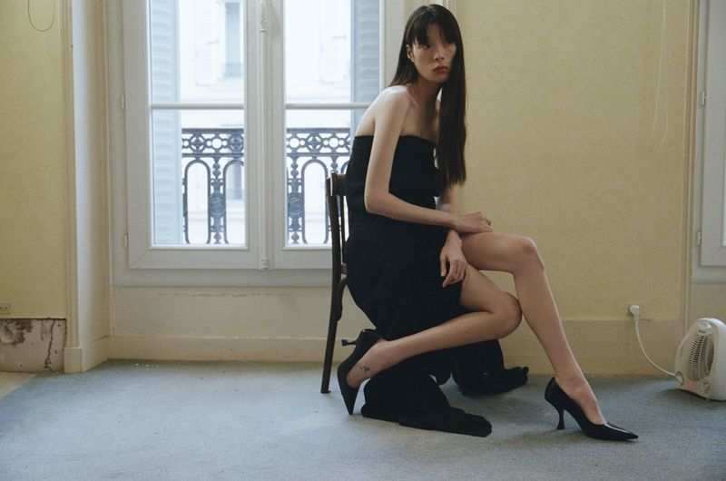 A slender woman with long dark hair poses seated in a short strapless black dress and heels inside a room with large windows.