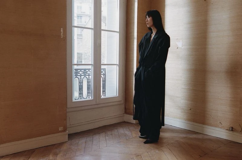 Indoor portrait of a slender adult woman in a dark coat standing near a window with textured walls and wooden floor, eye-level angle.