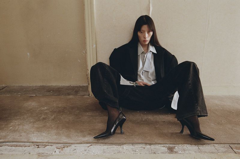 Model with long dark hair seated on the floor wearing an oversized black blazer, white collared shirt, wide trousers, and pointed heels against a neutral wall.