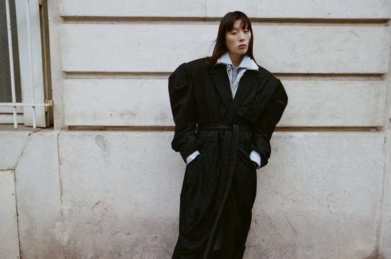 A full-length shot of an Asian woman with long dark hair wearing a long black coat with large sleeves posing outdoors.