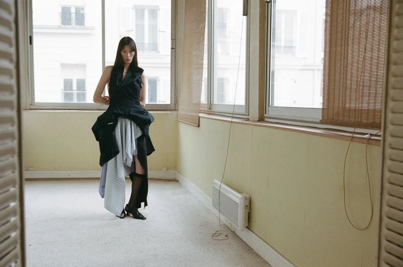 A slender woman in avant-garde black dress stands in a bright, empty room with large windows, showcasing a unique fashion style.