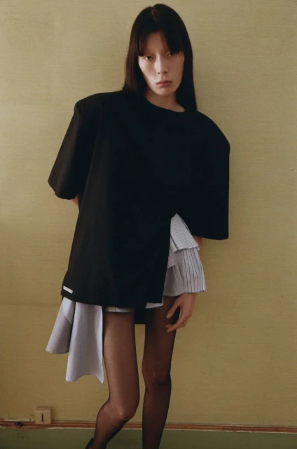 A single young adult female model poses in a stylish black top and light blue skirt with dark tights in front of a tan background.