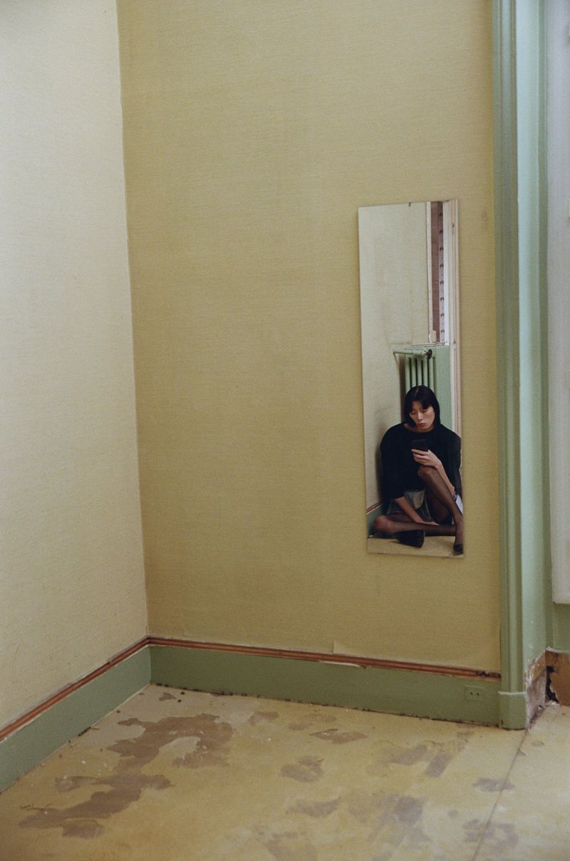 Woman sitting against a yellow wall reflected in a mirror. The room is sparsely furnished with a radiator and damaged floor.