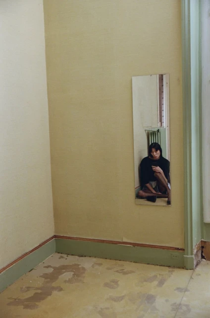 Woman sitting against a yellow wall reflected in a mirror. The room is sparsely furnished with a radiator and damaged floor.