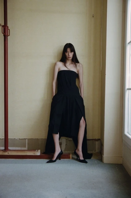 Elegant adult female fashion model in a long black strapless dress and heeled shoes posing indoors against a faded yellow wall.