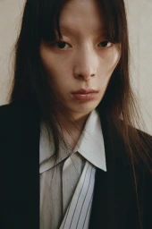 Close-up portrait of an East Asian woman wearing a white collared shirt and black blazer against a plain wall.