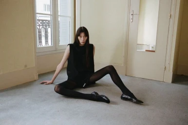 Model seated on the floor wearing a black top and tights against an interior room background