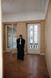 Model wearing an oversized dark coat standing in an empty room with herringbone parquet floors and tall windows.