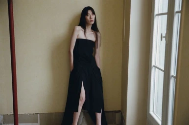 Model in a long black strapless dress with a high slit leaning against a pale yellow wall by a window