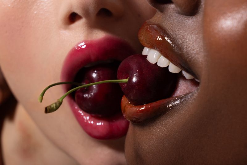 Close-up of two faces with open mouths sharing cherries, showcasing glossy lips, perfect teeth, and striking diversity.