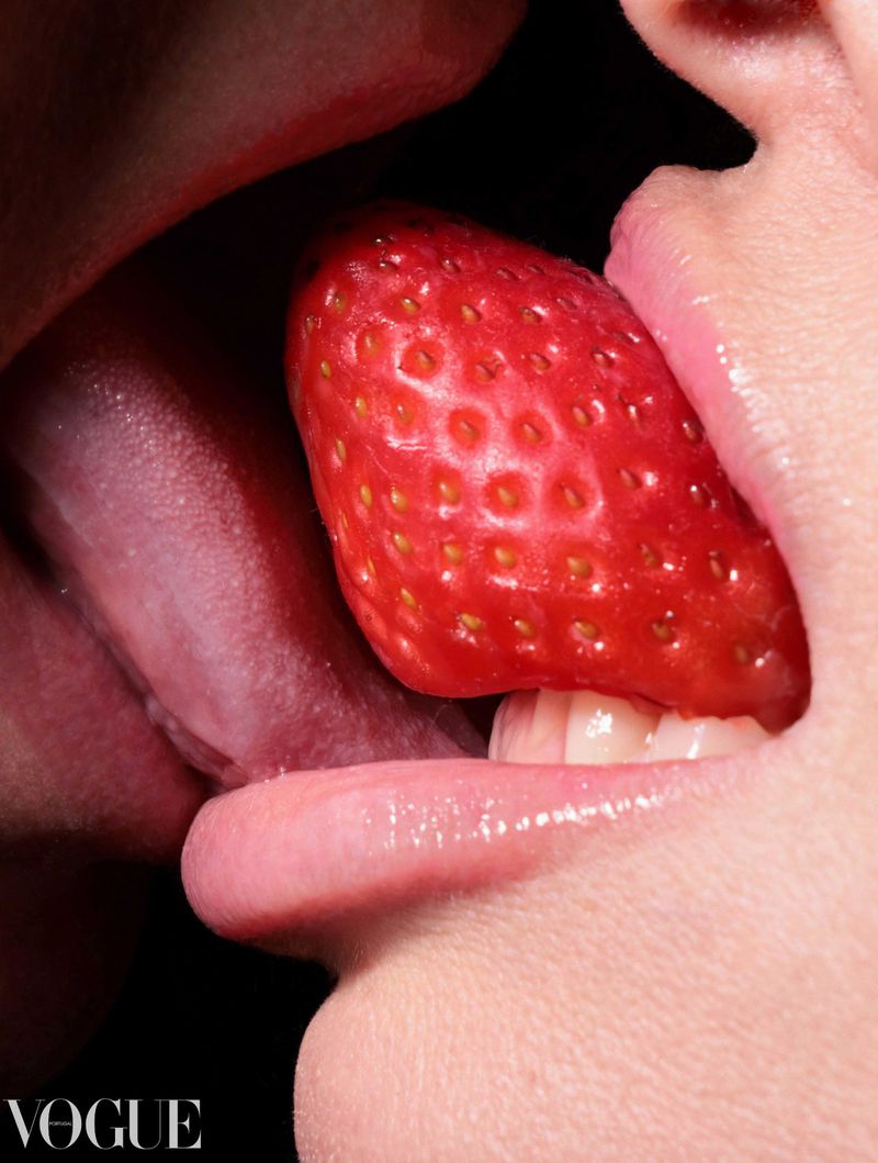 Close-up of two faces sharing a ripe red strawberry, lips touching, in a suggestive image against a dark background.
