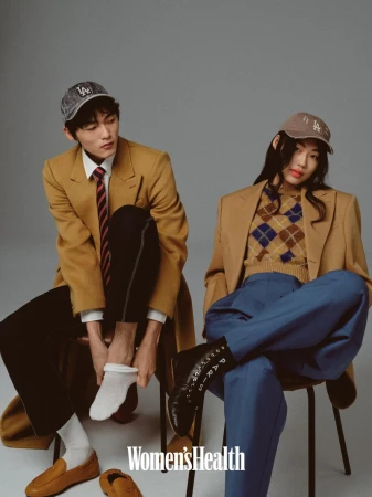 Man and woman seated on chairs wearing camel blazers, baseball caps, and matching styled attire against a plain grey backdrop.