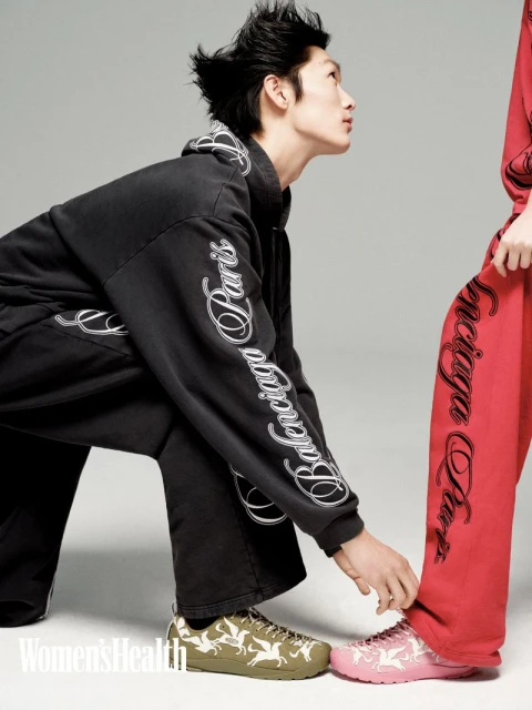 An Asian male model in a black Balenciaga tracksuit kneeling next to a woman in a red tracksuit, both wearing printed sneakers, on a white background.