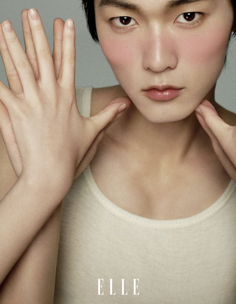 Close-up studio shot of an Asian male model with blush, hands framing his face, and wearing a white tank top, plus Elle logo.