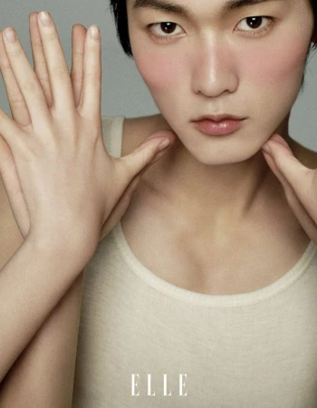 Close-up studio shot of an Asian male model with blush, hands framing his face, and wearing a white tank top, plus Elle logo.