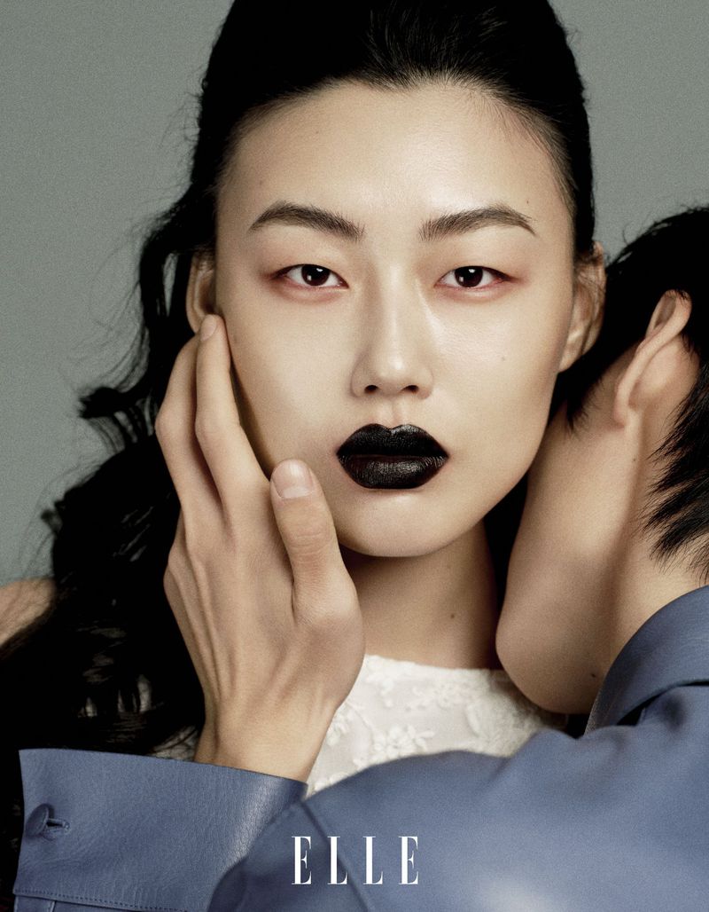 Close-up fashion shot of an Asian woman with black lipstick and dark hair, alongside a second person wearing a blue jacket. Elle logo is visible.