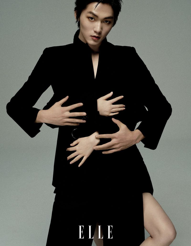 A person in a black blazer and skirt poses with arms and hands across their torso and leg. ELLE brand logo in the corner.