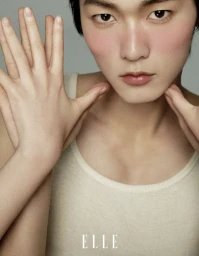 Close-up portrait of an East Asian male model holding his hands up to his face wearing a cream tank top against a grey background.