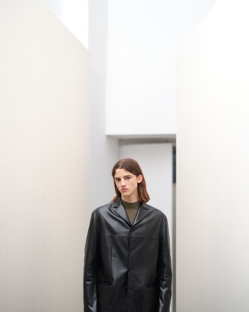 Portrait of an adult male with long brown hair wearing a black leather coat standing in a white interior corridor.