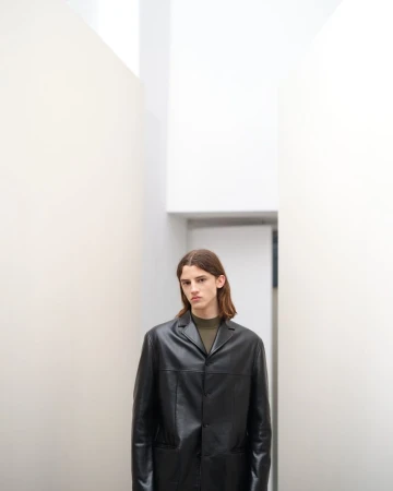 Portrait of an adult male with long brown hair wearing a black leather coat standing in a white interior corridor.