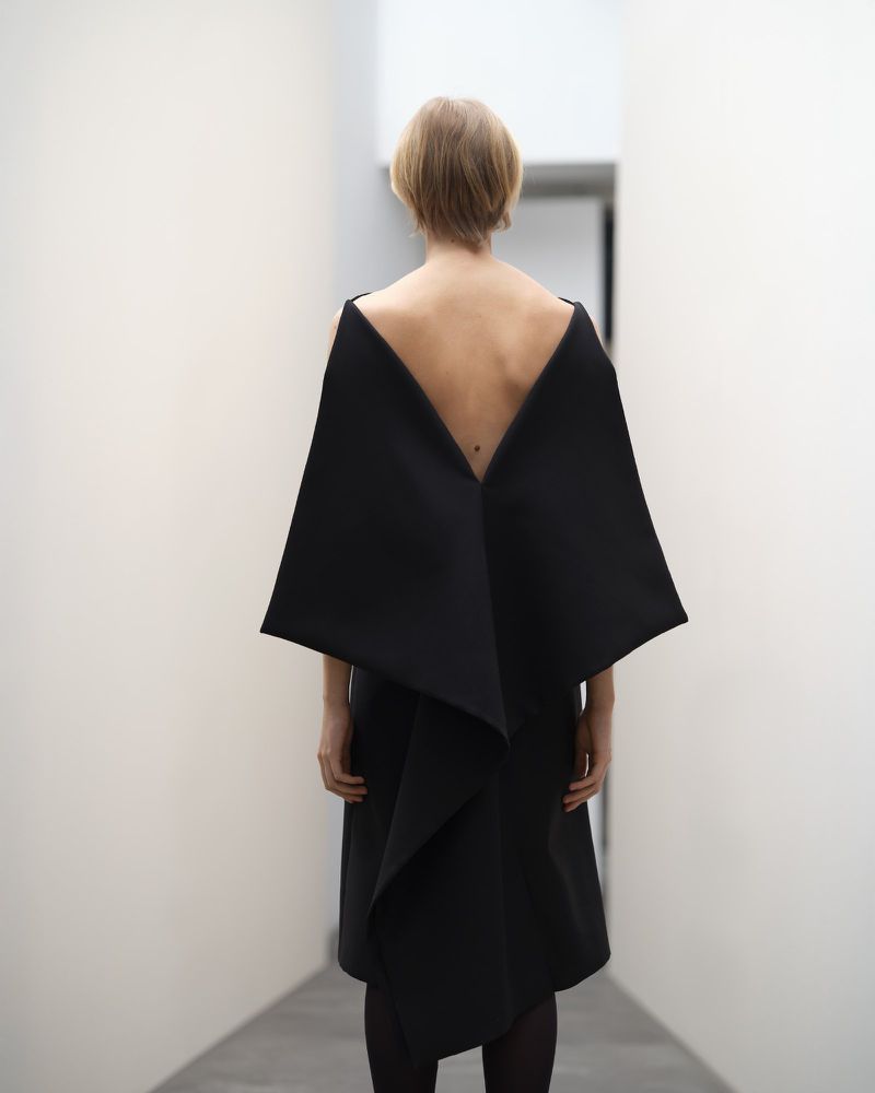 Woman in a black dress with an open back stands in a bright white hallway, creating a minimalist and modern aesthetic.