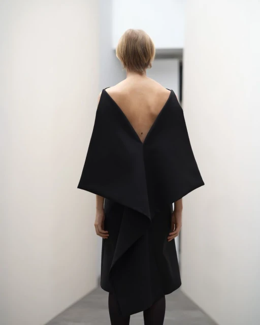 Woman in a black dress with an open back stands in a bright white hallway, creating a minimalist and modern aesthetic.