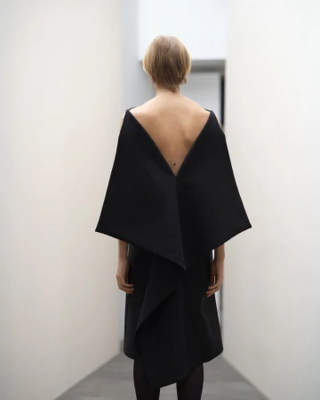 Woman in a black dress with an open back stands in a bright white hallway, creating a minimalist and modern aesthetic.