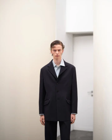 Male model in a dark blazer and light button-down shirt standing in a minimalist interior space.