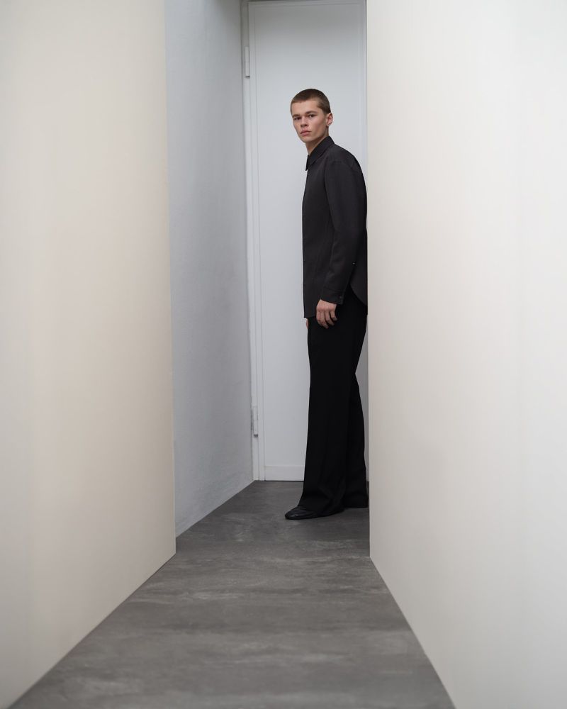 A young adult man in dark clothing stands between white walls in a brightly lit indoor setting, looking toward the camera.