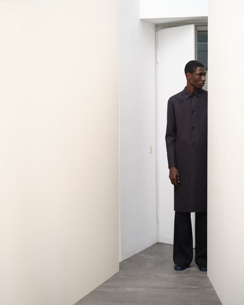 An African American male model in a dark overcoat and trousers stands between white walls in a bright interior space, fashion shoot.