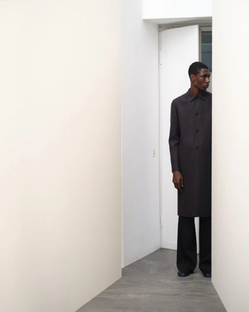 An African American male model in a dark overcoat and trousers stands between white walls in a bright interior space, fashion shoot.