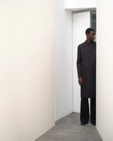 An African American male model in a dark overcoat and trousers stands between white walls in a bright interior space, fashion shoot.