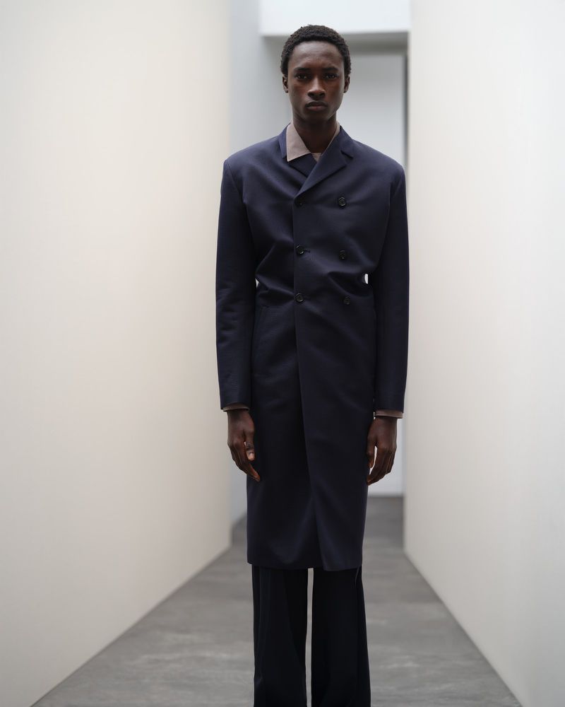 Man in a long navy coat and black pants stands in an illuminated hallway, fashion portrait with clean studio aesthetic.