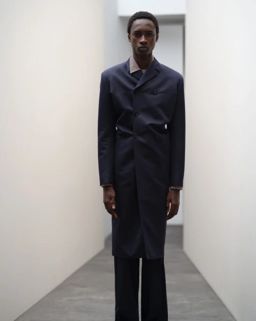 Man in a long navy coat and black pants stands in an illuminated hallway, fashion portrait with clean studio aesthetic.