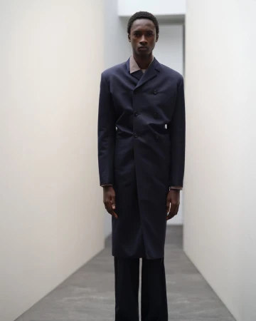Man in a long navy coat and black pants stands in an illuminated hallway, fashion portrait with clean studio aesthetic.
