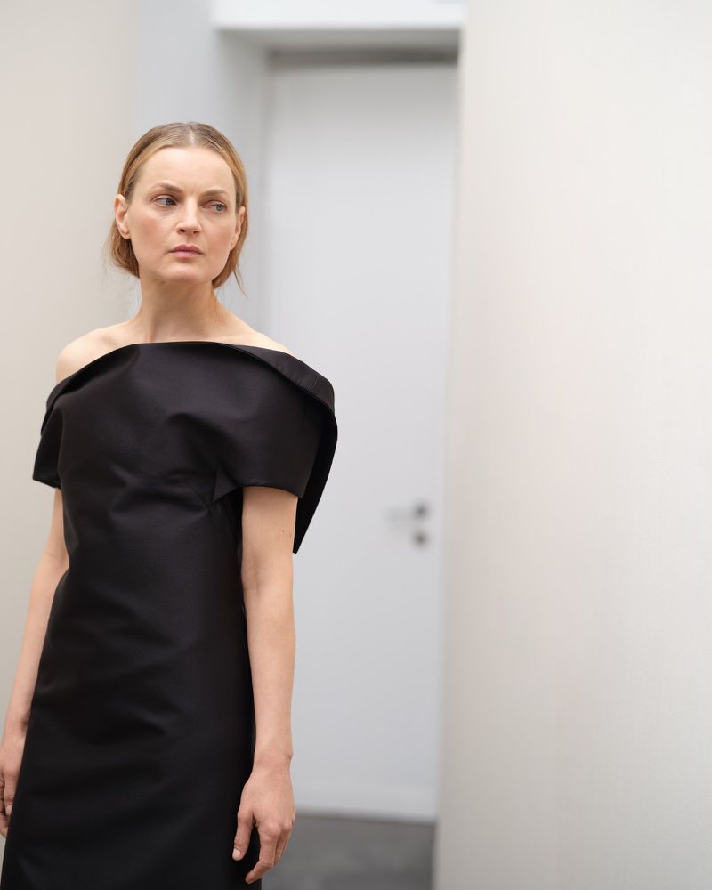 Woman in black off-the-shoulder dress standing in a bright, neutral room, looking away with a contemplative expression.