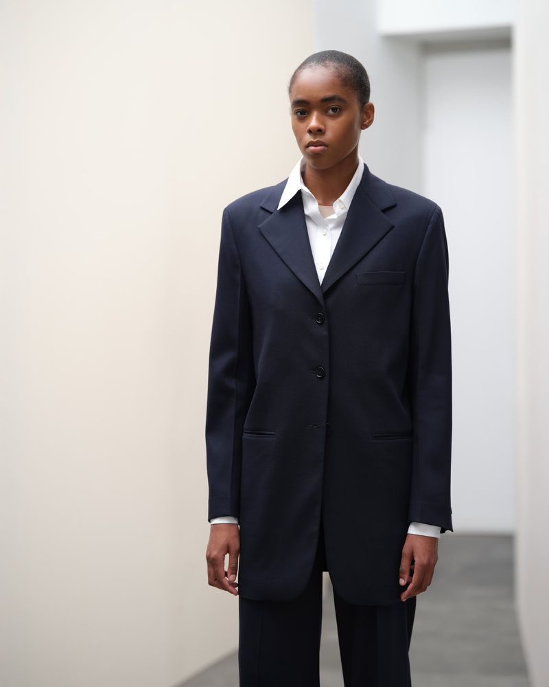 Portrait of a black woman in a dark blue suit and white shirt posing against a plain indoor backdrop with neutral colors.