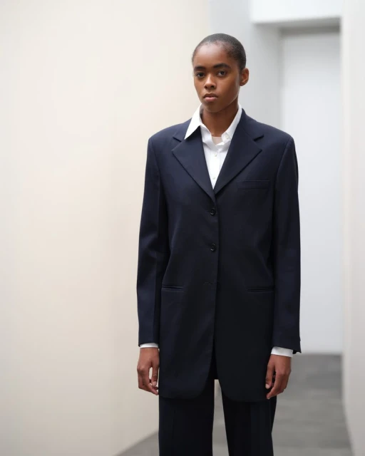 Portrait of a black woman in a dark blue suit and white shirt posing against a plain indoor backdrop with neutral colors.