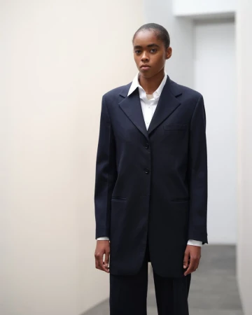 Portrait of a black woman in a dark blue suit and white shirt posing against a plain indoor backdrop with neutral colors.