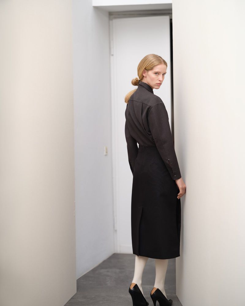 Blonde adult female in dark top and black skirt stands in white hallway with geometric heels and opaque tights looking at camera.