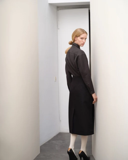 Blonde adult female in dark top and black skirt stands in white hallway with geometric heels and opaque tights looking at camera.