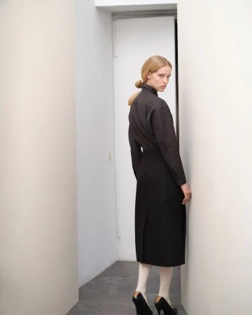 Blonde adult female in dark top and black skirt stands in white hallway with geometric heels and opaque tights looking at camera.