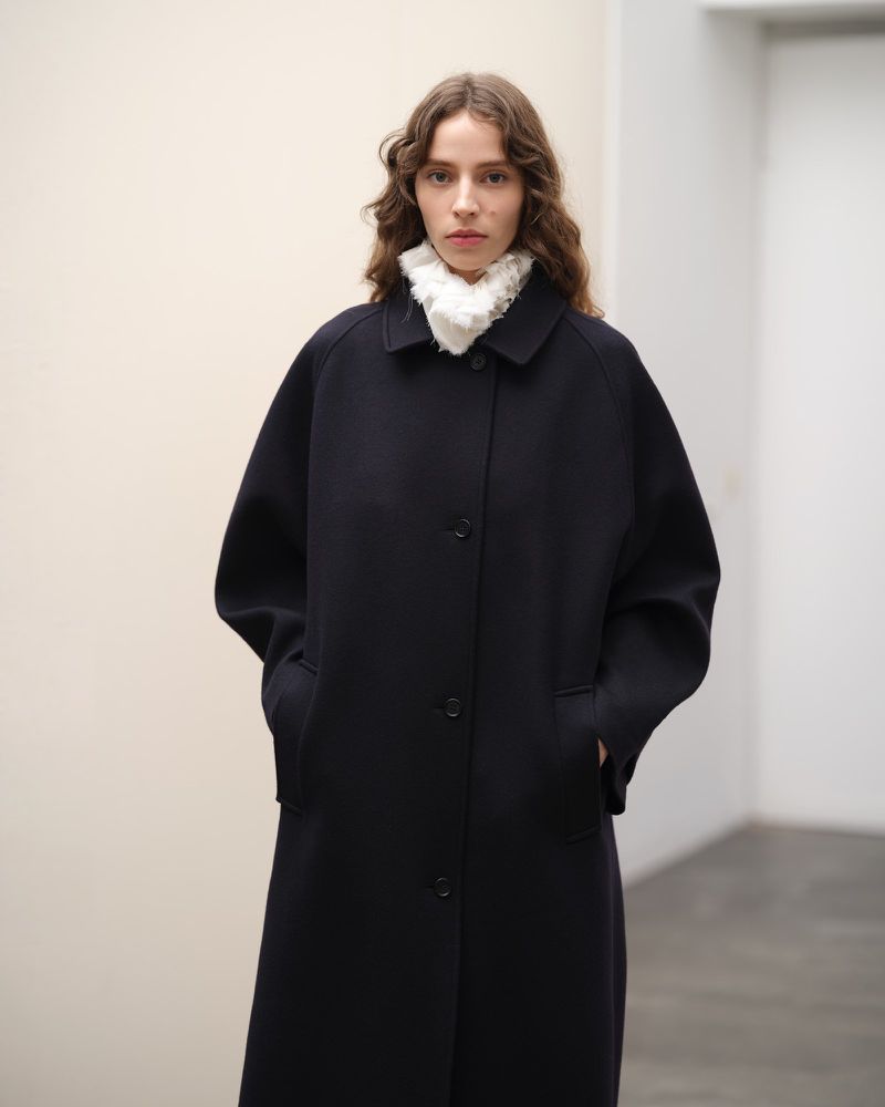 A woman poses indoors, wearing a long navy coat and white ruffled collar, hands in pockets, standing in front of a neutral background.
