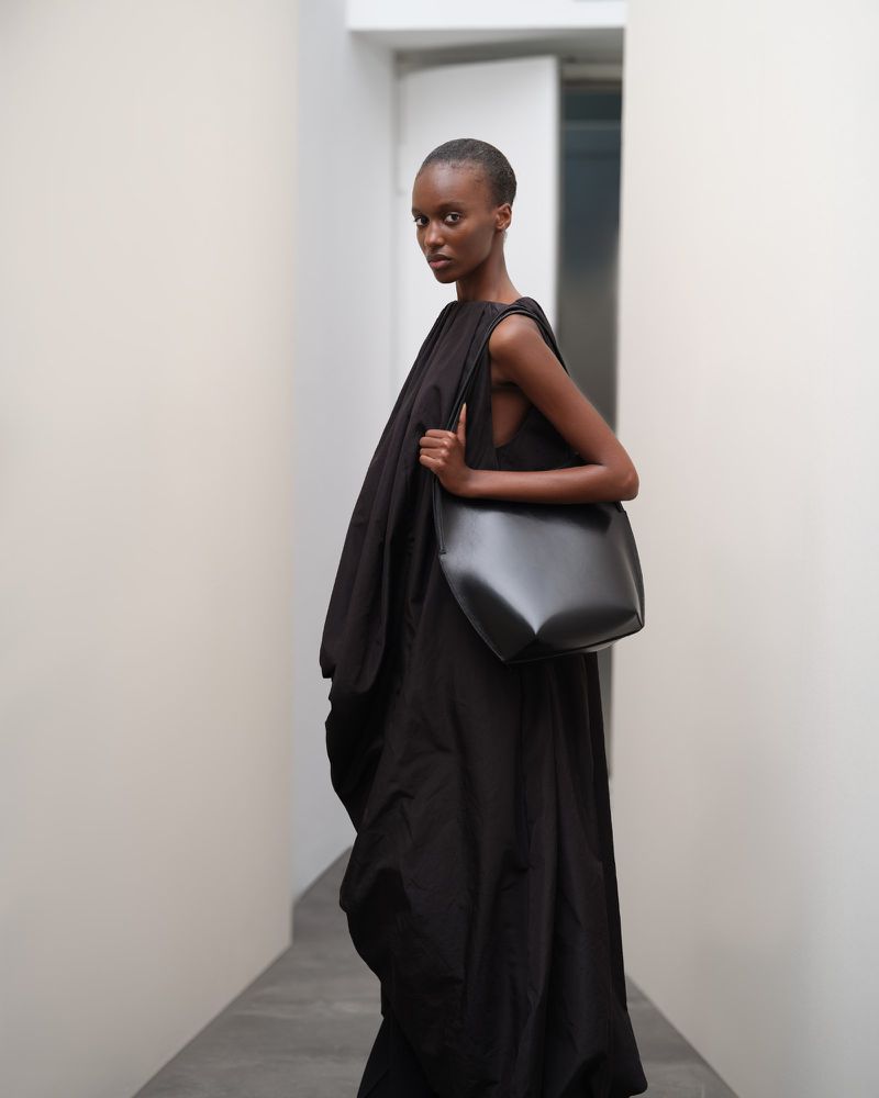 Fashion shot of a Black female model wearing a dark dress and carrying a dark leather bag in an indoor corridor setting.
