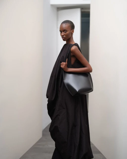 Fashion shot of a Black female model wearing a dark dress and carrying a dark leather bag in an indoor corridor setting.