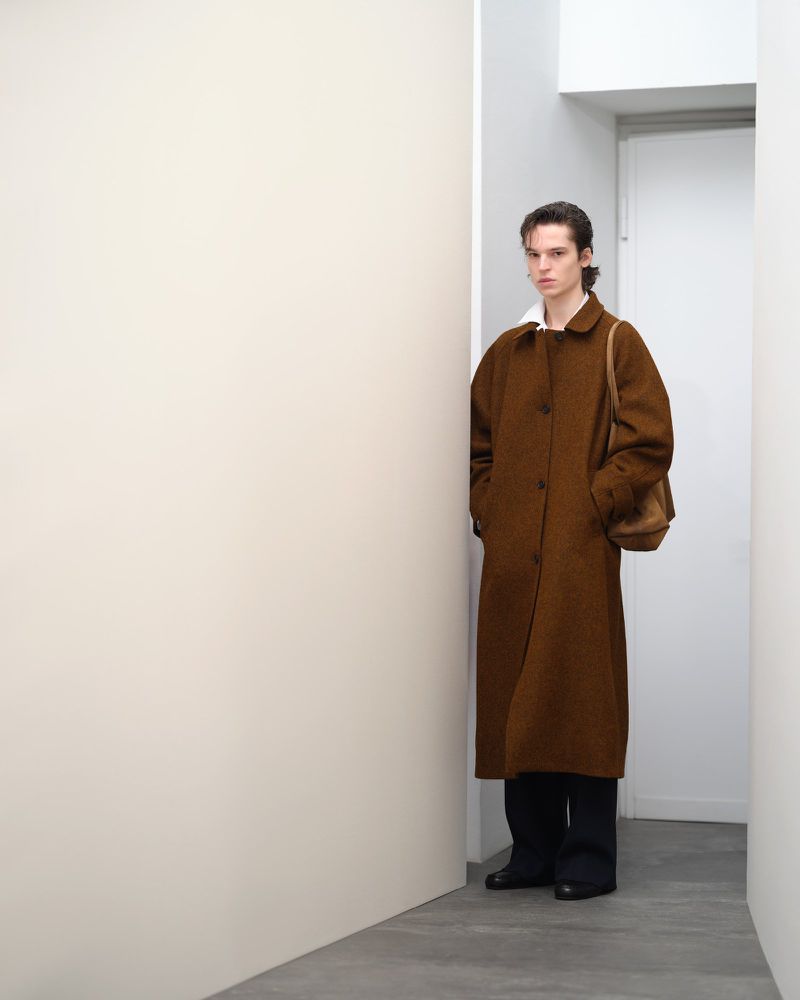 An adult male in a brown overcoat with a shoulder bag poses indoors against a white wall, creating a minimalist fashion editorial look.
