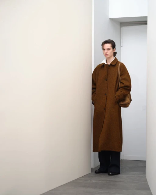 An adult male in a brown overcoat with a shoulder bag poses indoors against a white wall, creating a minimalist fashion editorial look.