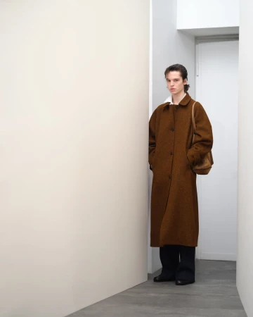 An adult male in a brown overcoat with a shoulder bag poses indoors against a white wall, creating a minimalist fashion editorial look.