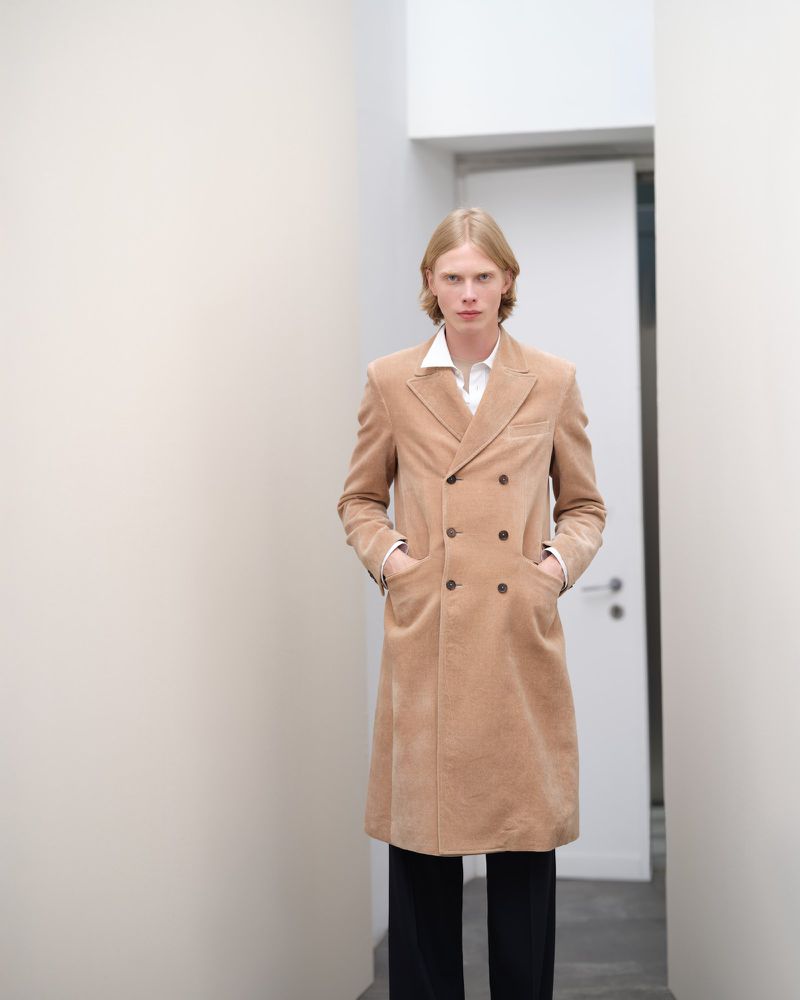 A blonde male model wears a tan corduroy double-breasted coat over a white shirt, standing in a minimalist white-walled corridor.