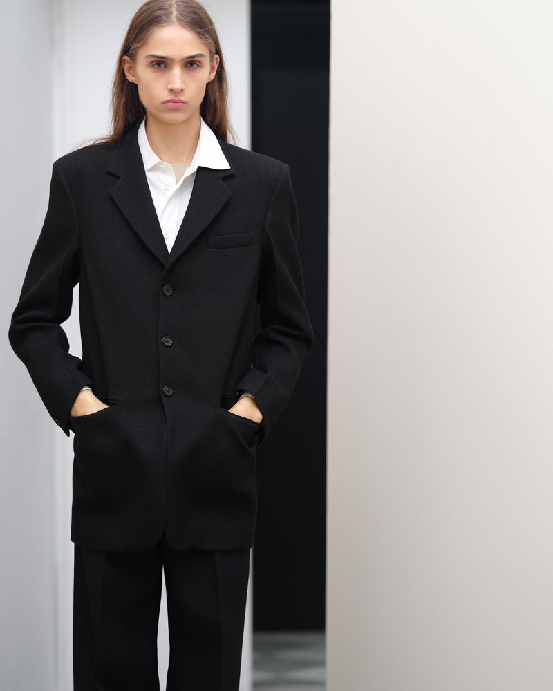 A young adult woman wearing a black suit with a white shirt poses indoors against a plain wall in a portrait-style fashion shot.